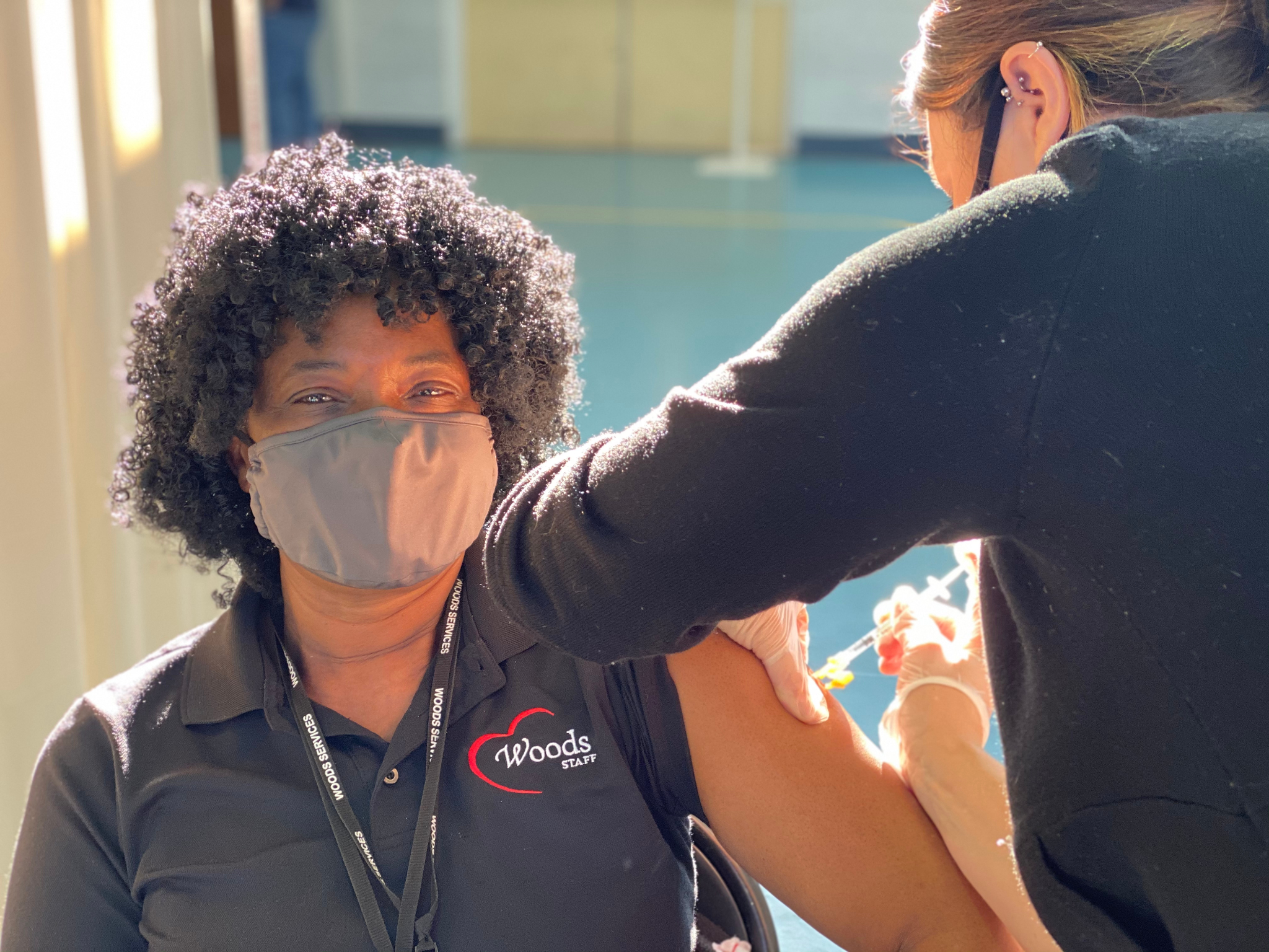 An employee of Woods Services with curly hair, wearing a tan mask, receives the first COVID vaccine on Dec. 22, 2020